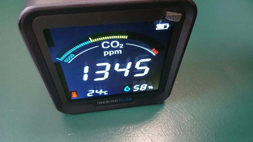 A carbon dioxide monitor sitting on a green pleather bench reads 1345 parts per million at 24 degrees Celsius and 58% relative humidity