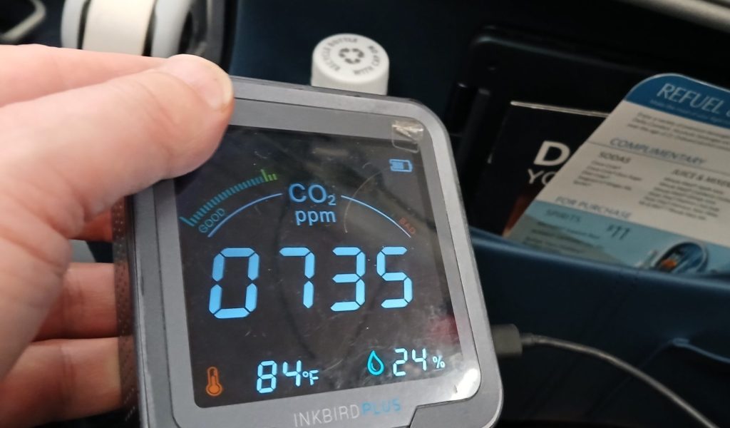 In an airplane seat, an INKBIRD carbon dioxide monitor reads 735 parts per million, 84 degrees Fahrenheit and 24% relative humidity.