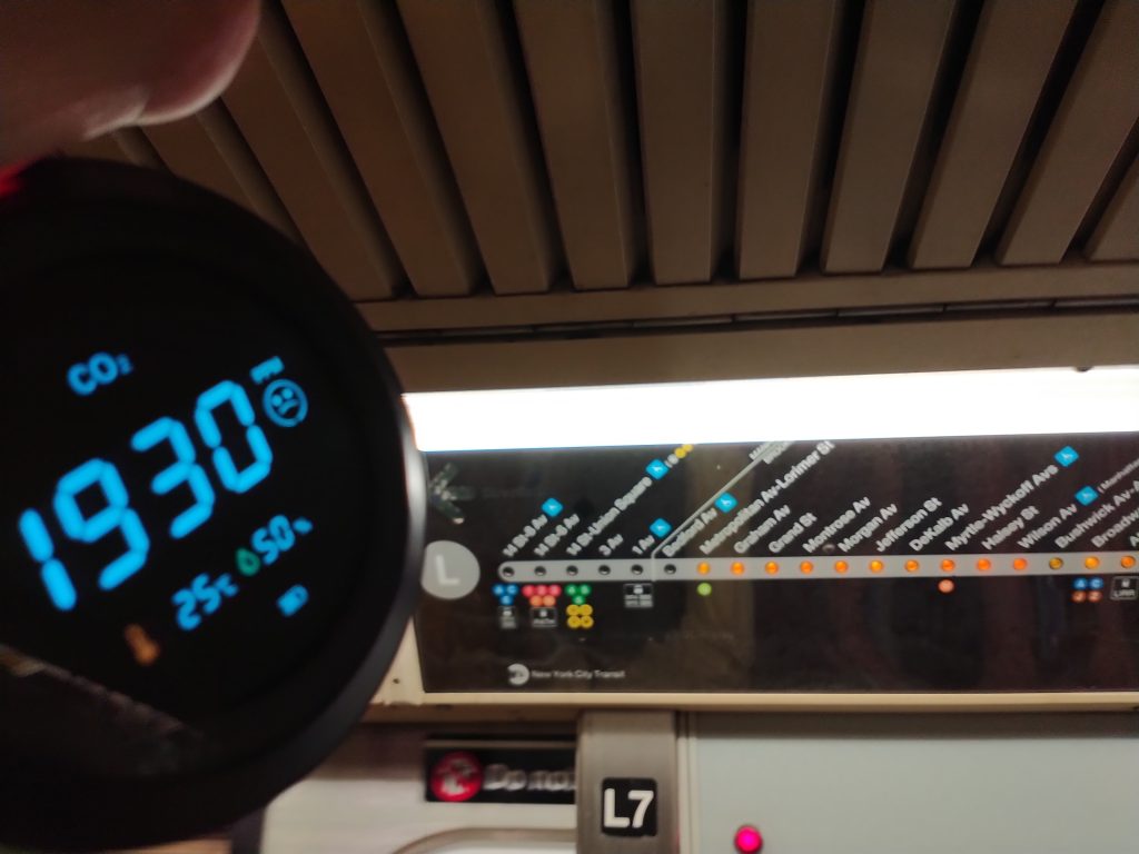 A carbon dioxide monitor reads 1930 parts per million, 25 degrees Celsius and 50% relative humidity. In the background is a strip map for the L train of the New York City subway, showing that the train is eastbound between Bedford Avenue and Lorimer Street.